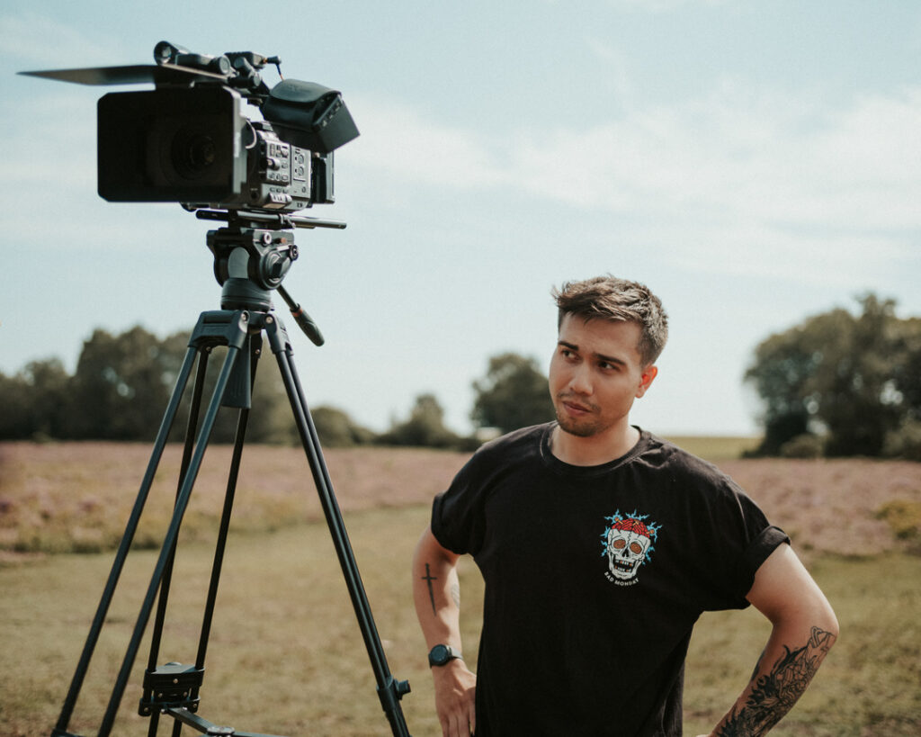 About Nathan Mountain Videography | A man stands outdoors with hands on his hips, looking at a video camera mounted on a tripod. He wears a black T-shirt with a colourful skull graphic, has tattoos on his left arm, and a watch on his right wrist. The background is a grassy field with trees under a blue sky. | Nathan Mountain | Freelance Camera Operator and Editor